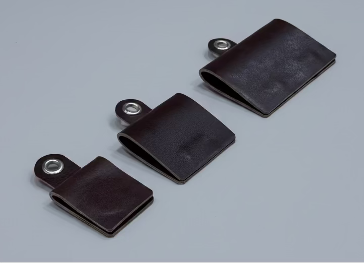 Three dark brown leather strap covers with metal snap loops, arranged diagonally on a light wood grain surface.